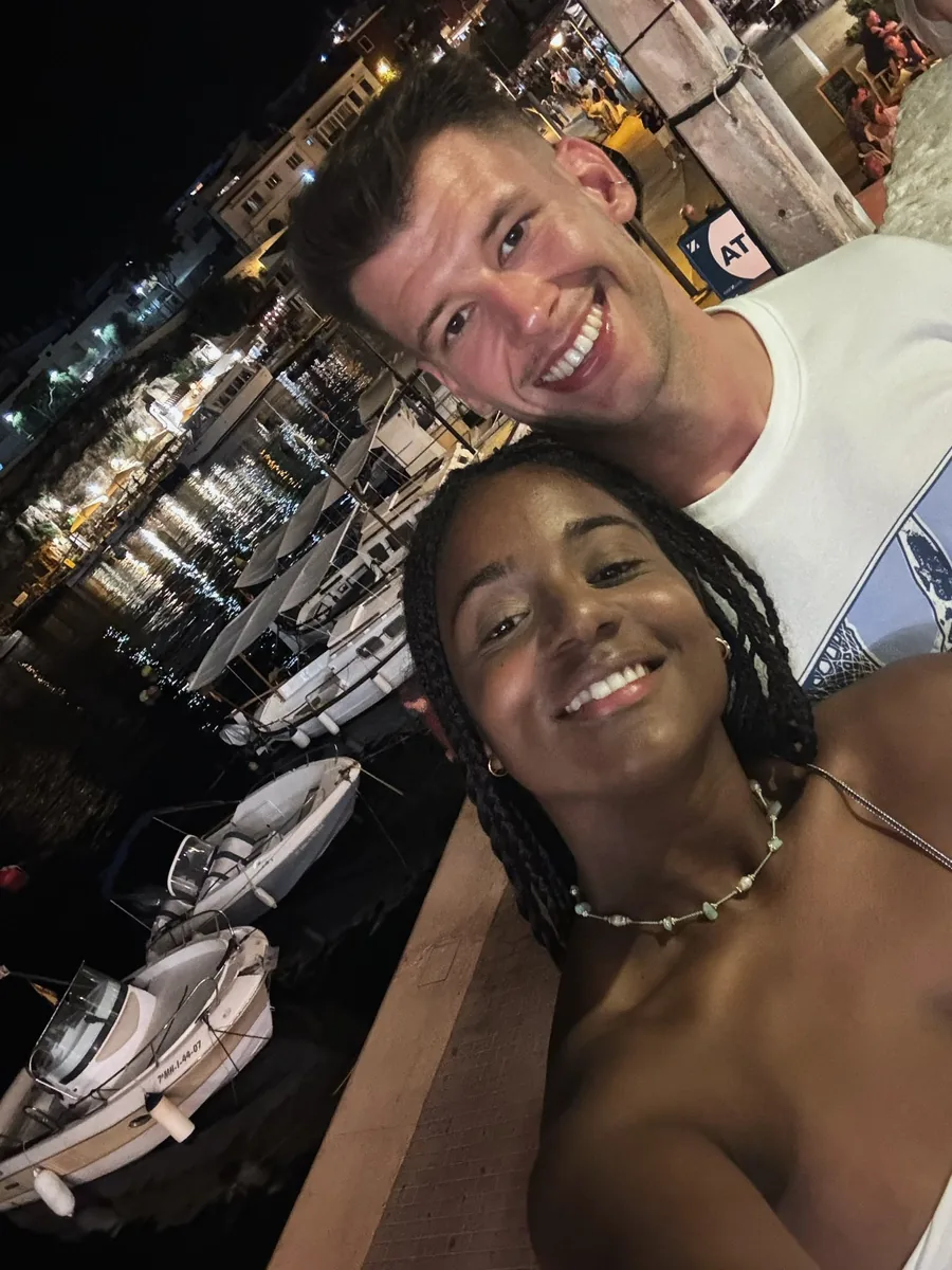Cinthia y Pablo de noche en el puerto, con los barcos amarrados y las luces del pueblo reflejadas en el agua